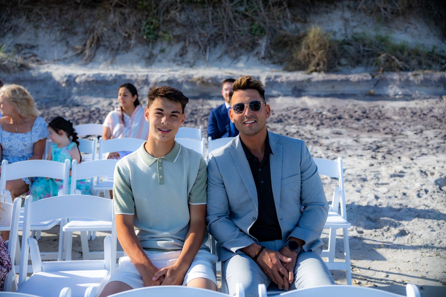 Two guests in smart casual attire sit on white chairs during an outdoor beach event.
