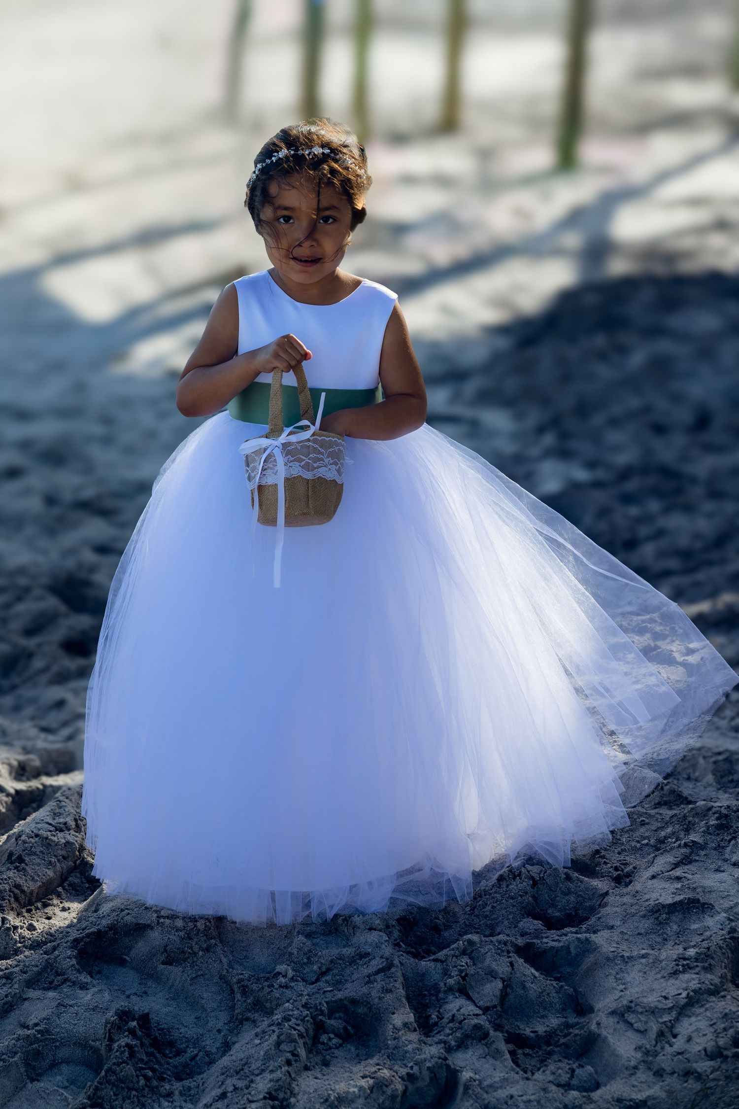 A child in a white tulle flower girl dress holding a small basket stands on sand at a beach wedding ceremony.