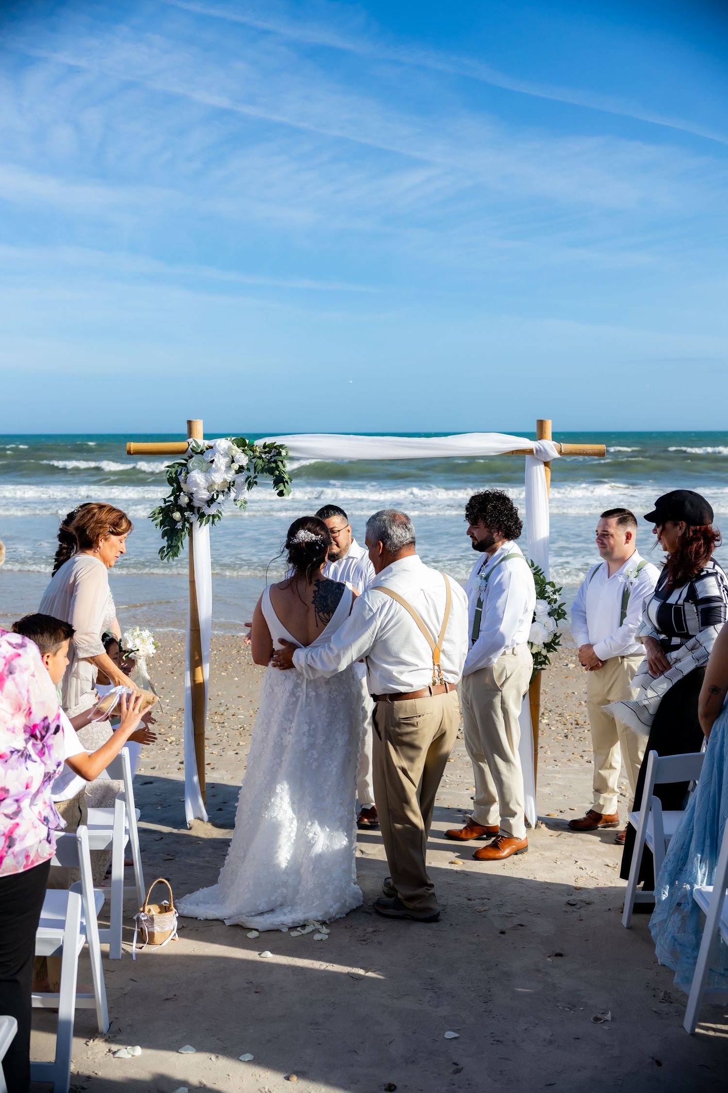 Romantic beach wedding ceremony under white arch with floral decorations on sandy shore by ocean.