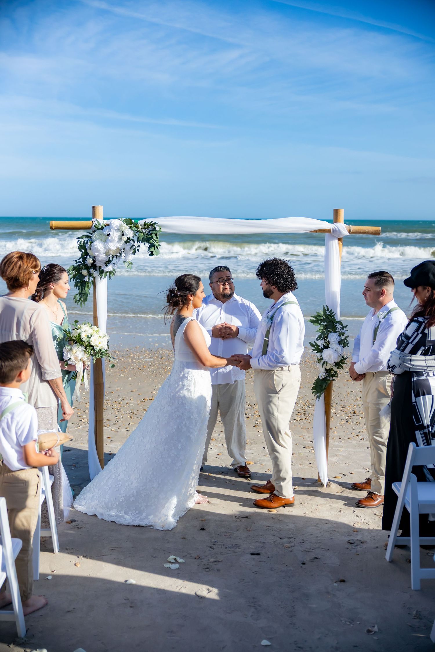 A beach wedding ceremony under a white arch decorated with flowers as waves crash in the background on a sunny day.