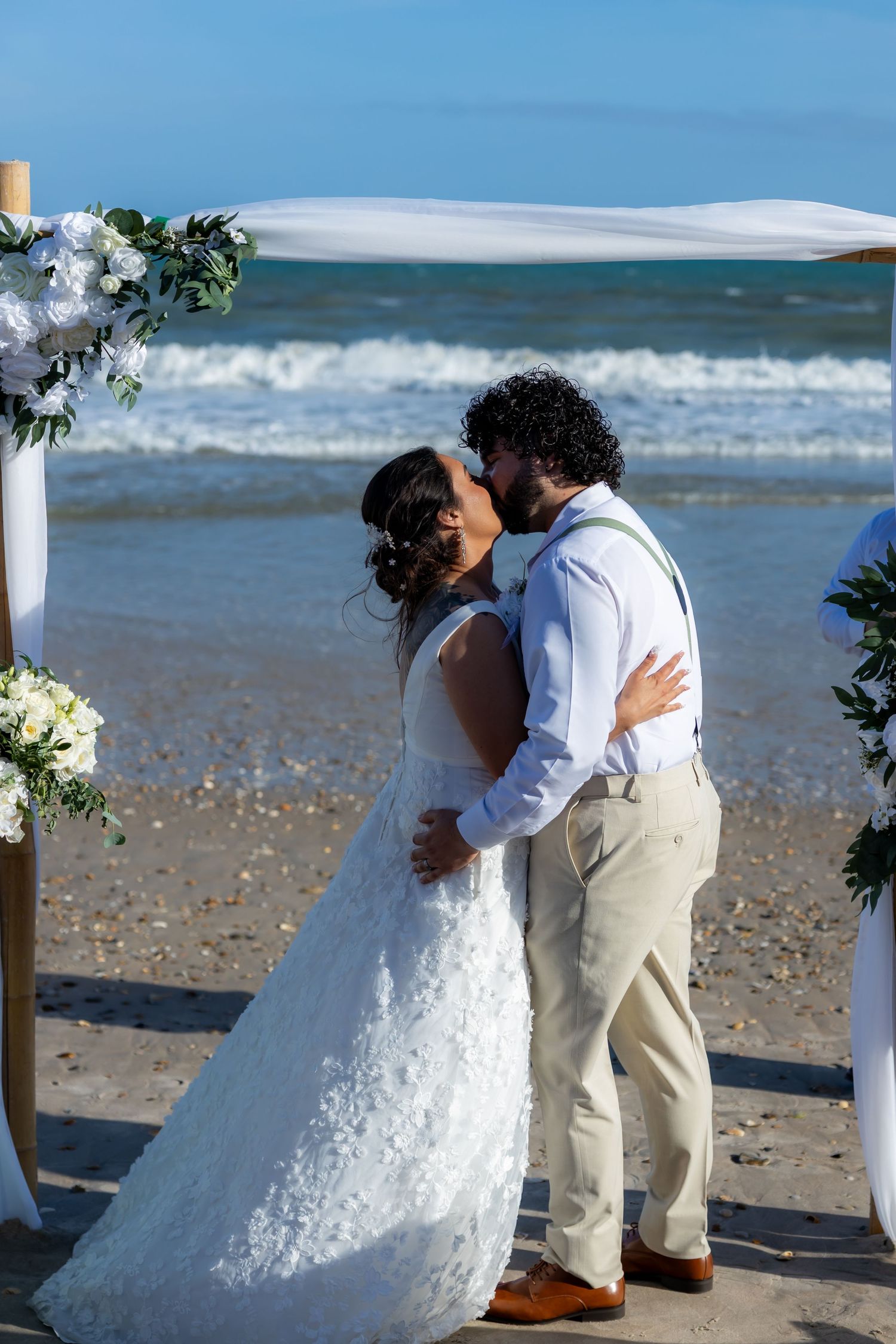 A couple shares their first kiss as newlyweds on a beach during a seaside wedding ceremony with white floral decor.