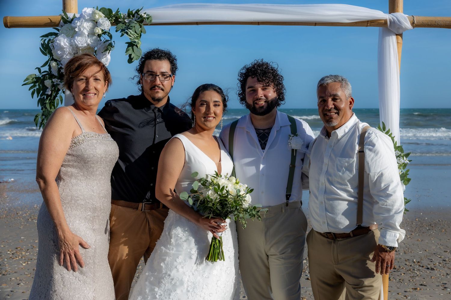 A wedding group poses together on a beach with the ocean in the background under a white fabric arch with greenery.