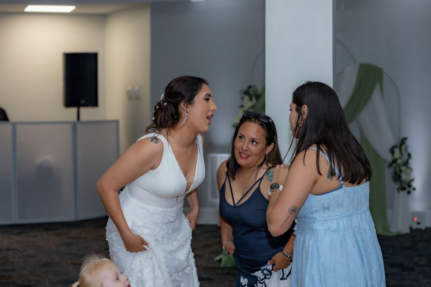 Three party guests share an animated conversation at an indoor event with one wearing a white dress and another in light blue.