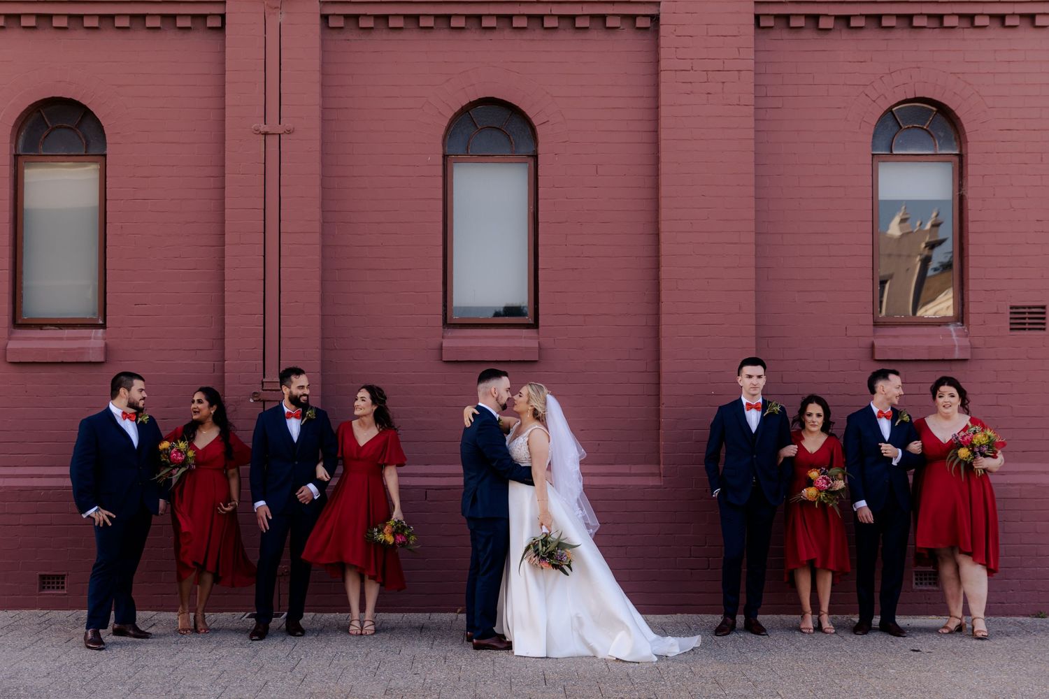 Wedding party stands in formal wear against red brick wall with arched windows during outdoor photo session.
