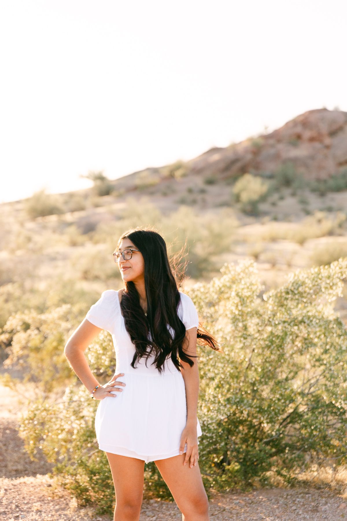 A woman in a white dress has a hand on her hip and is looking off. She has black hair and there's greenery and a mountain behind her.