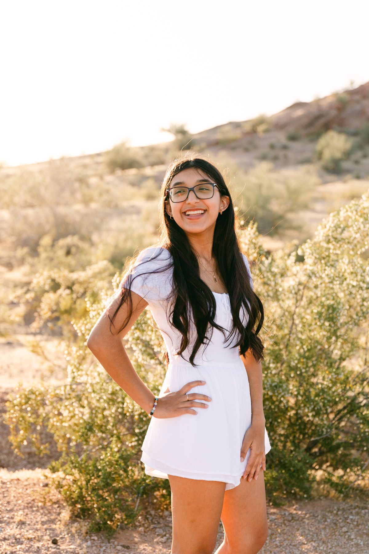 A woman in a white dress has a hand on her hip and is looking off. She has black hair and there's greenery and a mountain behind her.