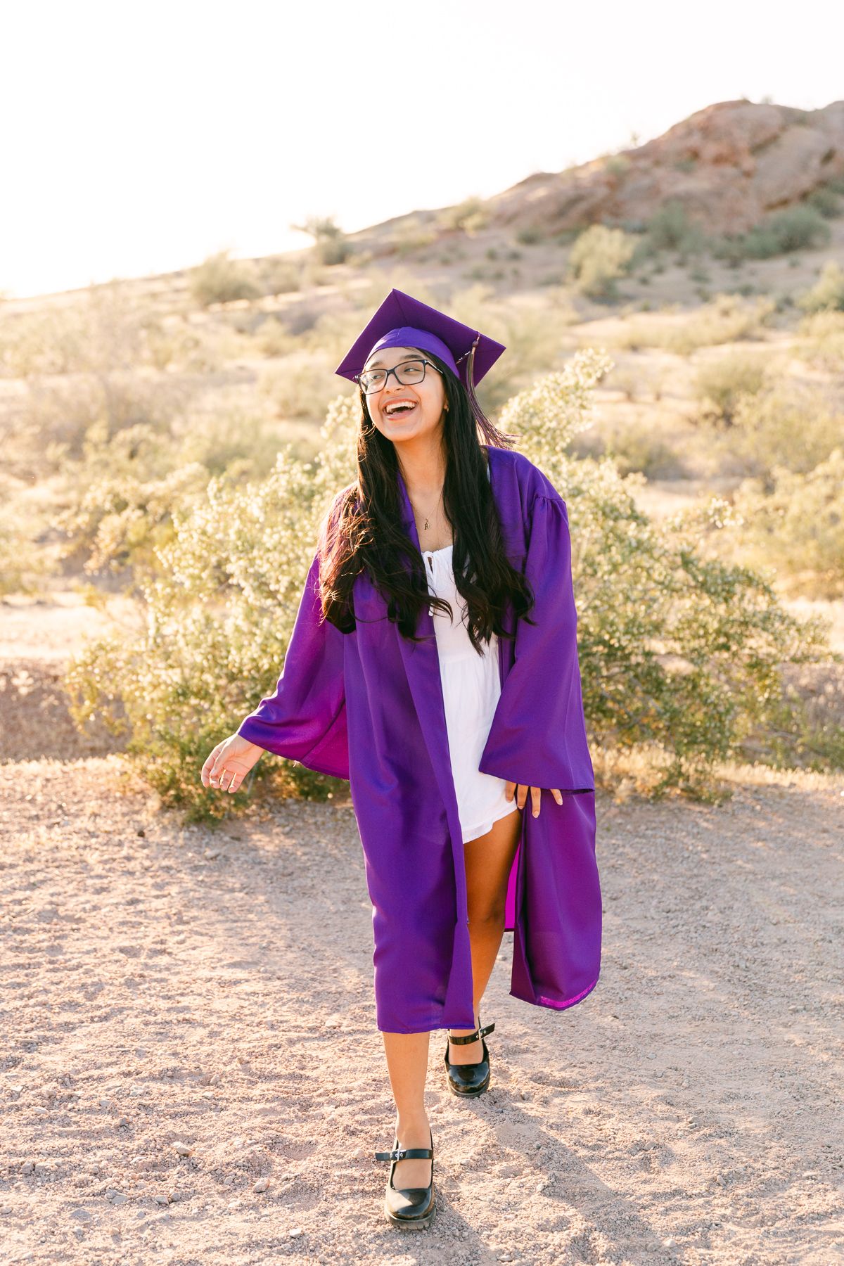 A woman is wearing a purple graduation gown and cap. She's smiling and walking forward. There's greenery and a mountain behind her.