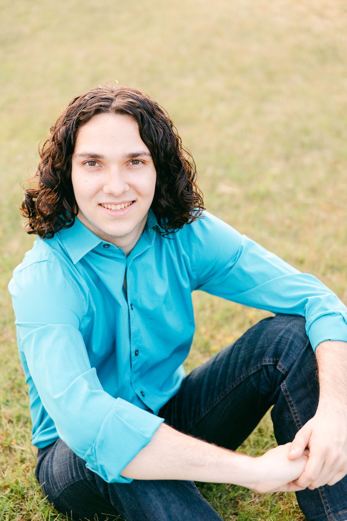 A man is sitting in the grass. He has brown curly hair, a blue shirt and jeans.