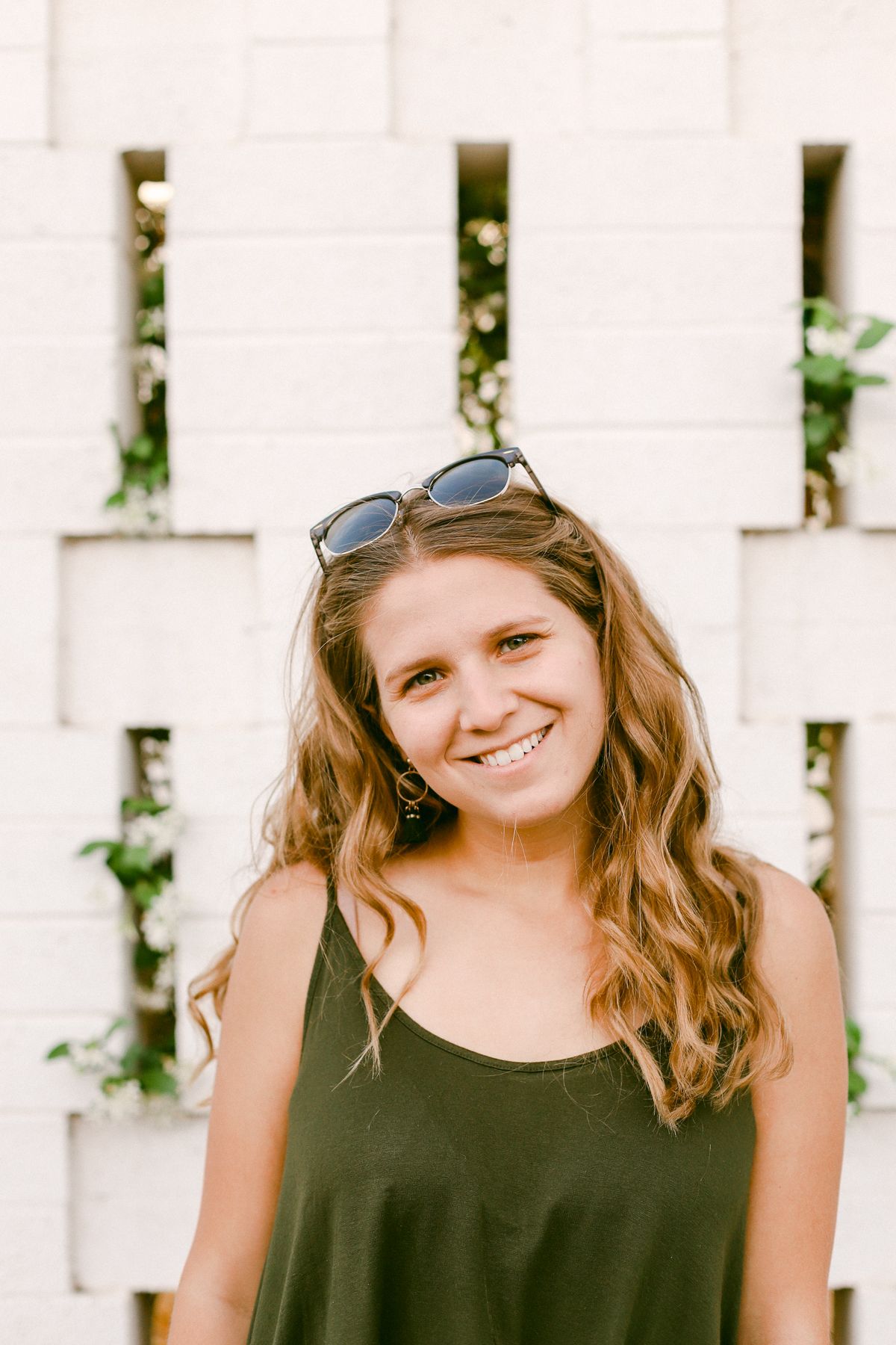 A woman with sunglasses and a green top. She's smiling and is standing in front of a white brick wall with greenery poking through