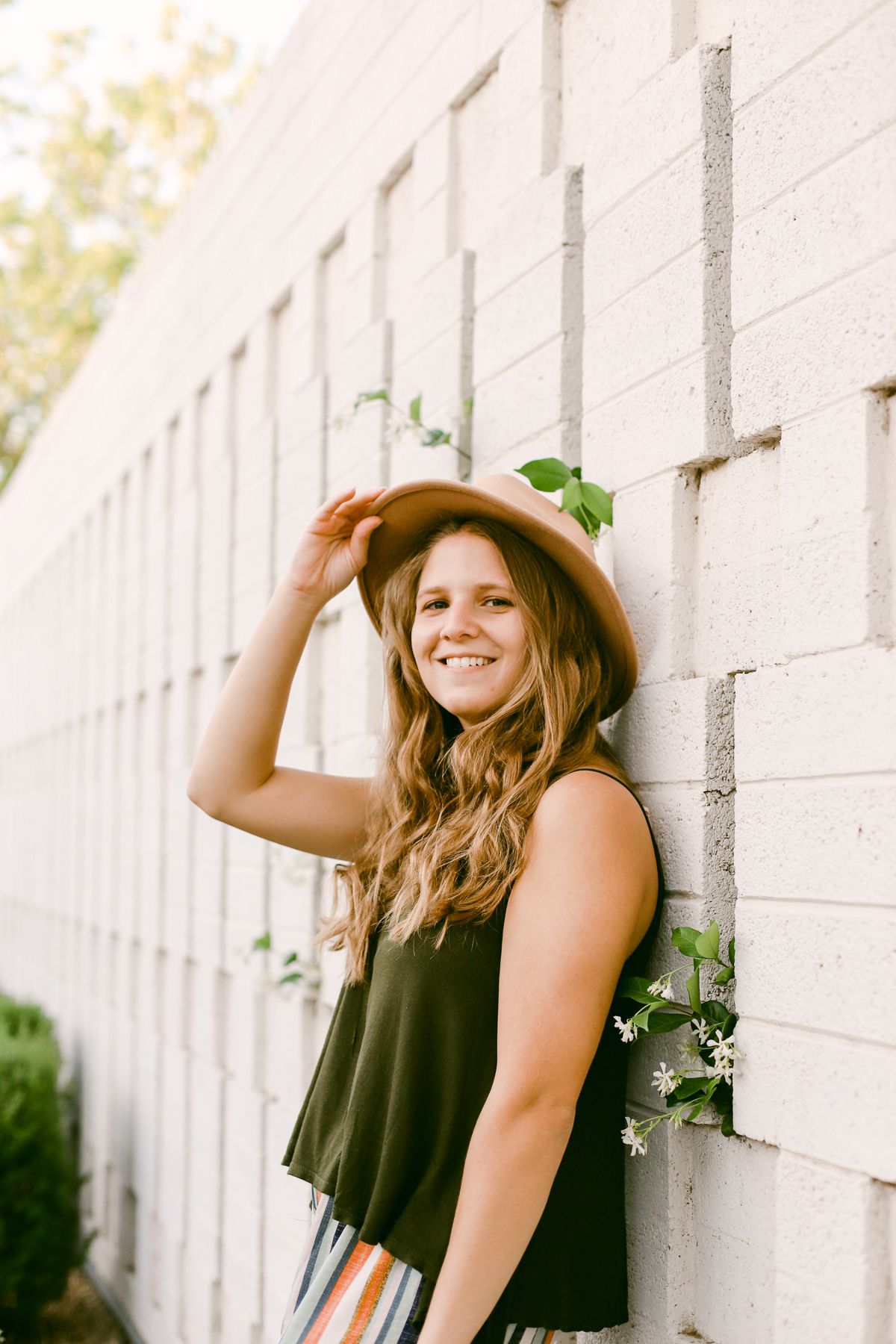 A woman in a green top with a brown hat. She's leaning against a white brick wall with greenery and has a hand on her hat.