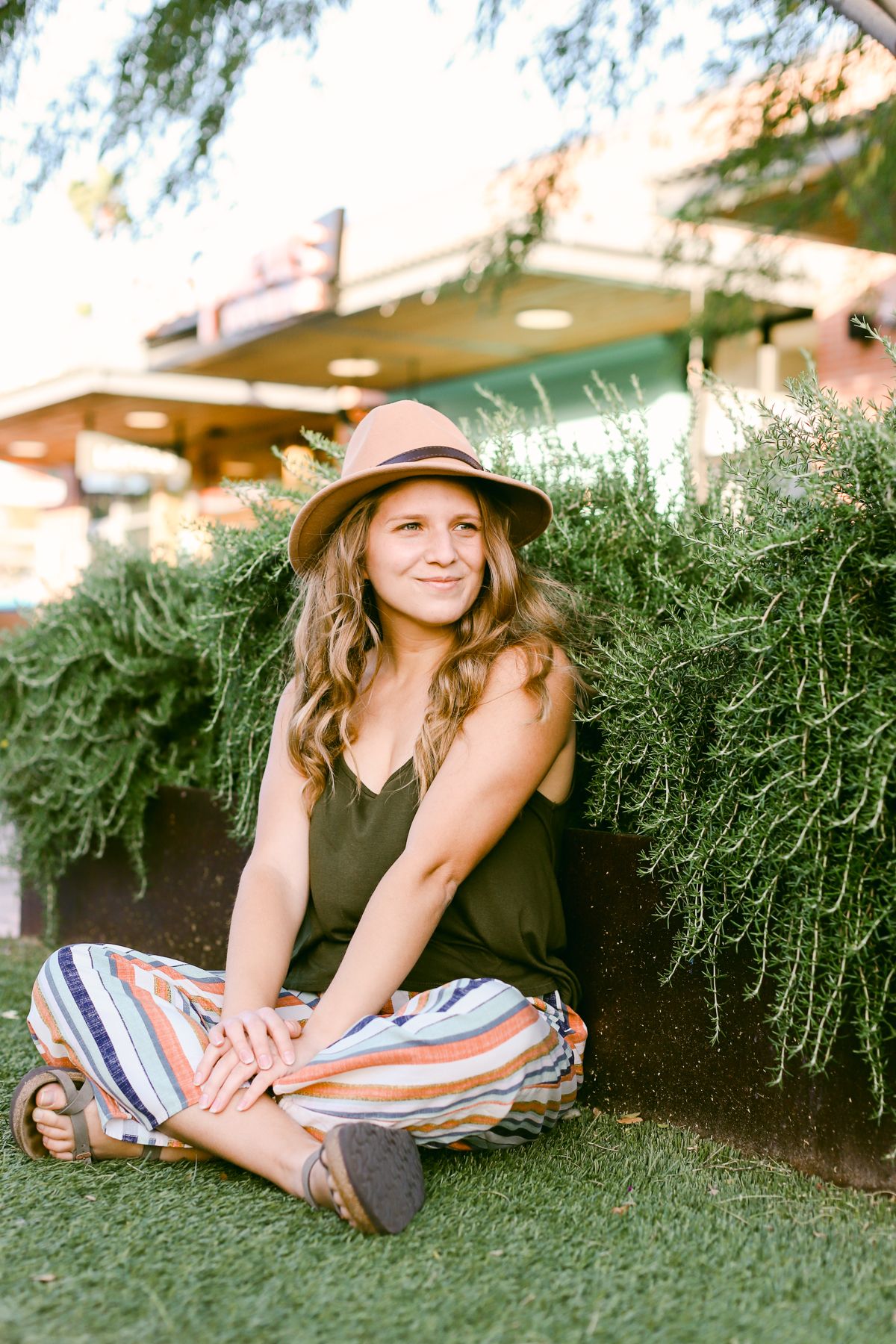 A smiling woman wearing a brown hat, green top and colorful pants. She's sitting on astroturf and has her back up against some greenery.