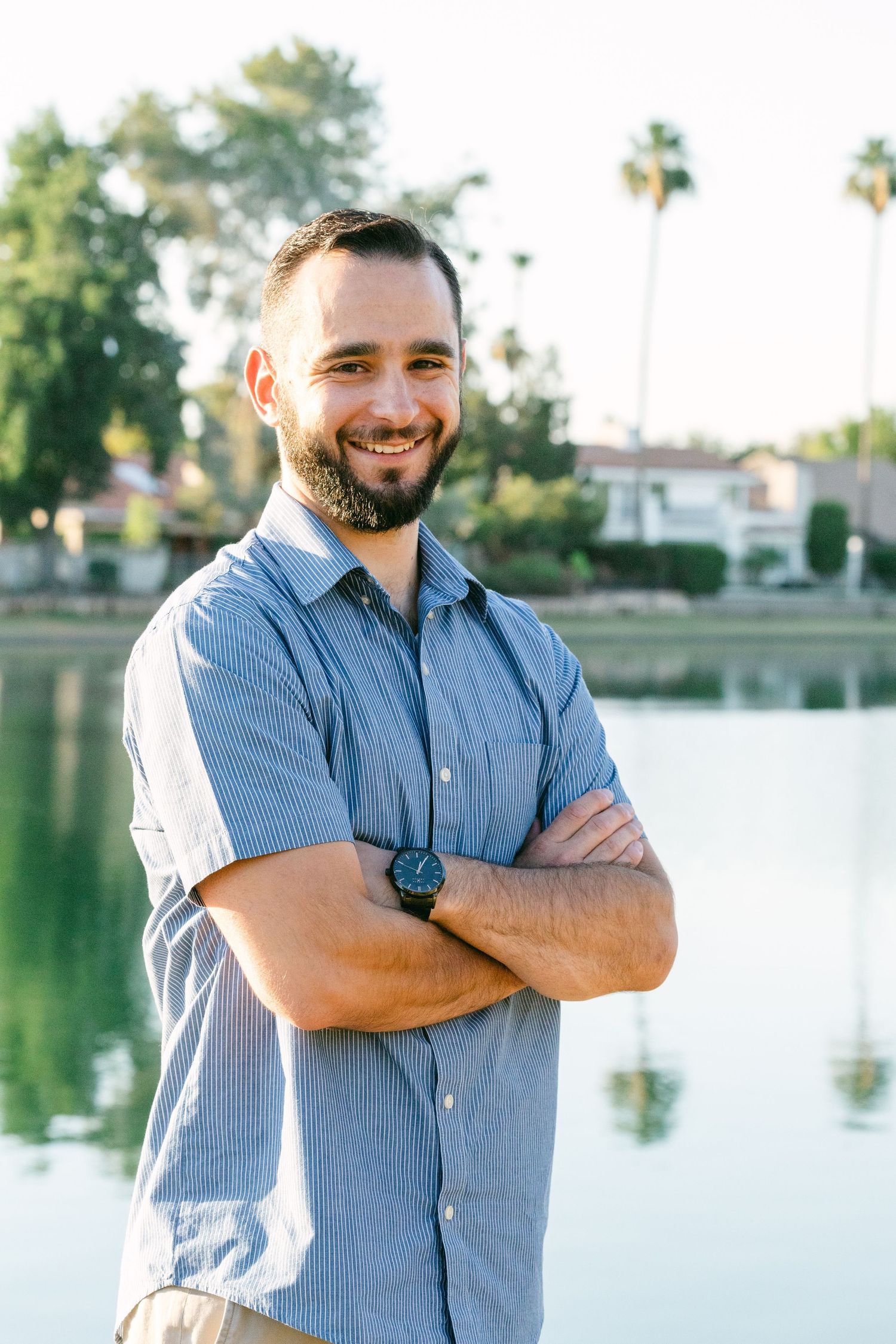 A man in a blue shirt has his arms crossed and is smiling. There's a lake and palm trees behind him.