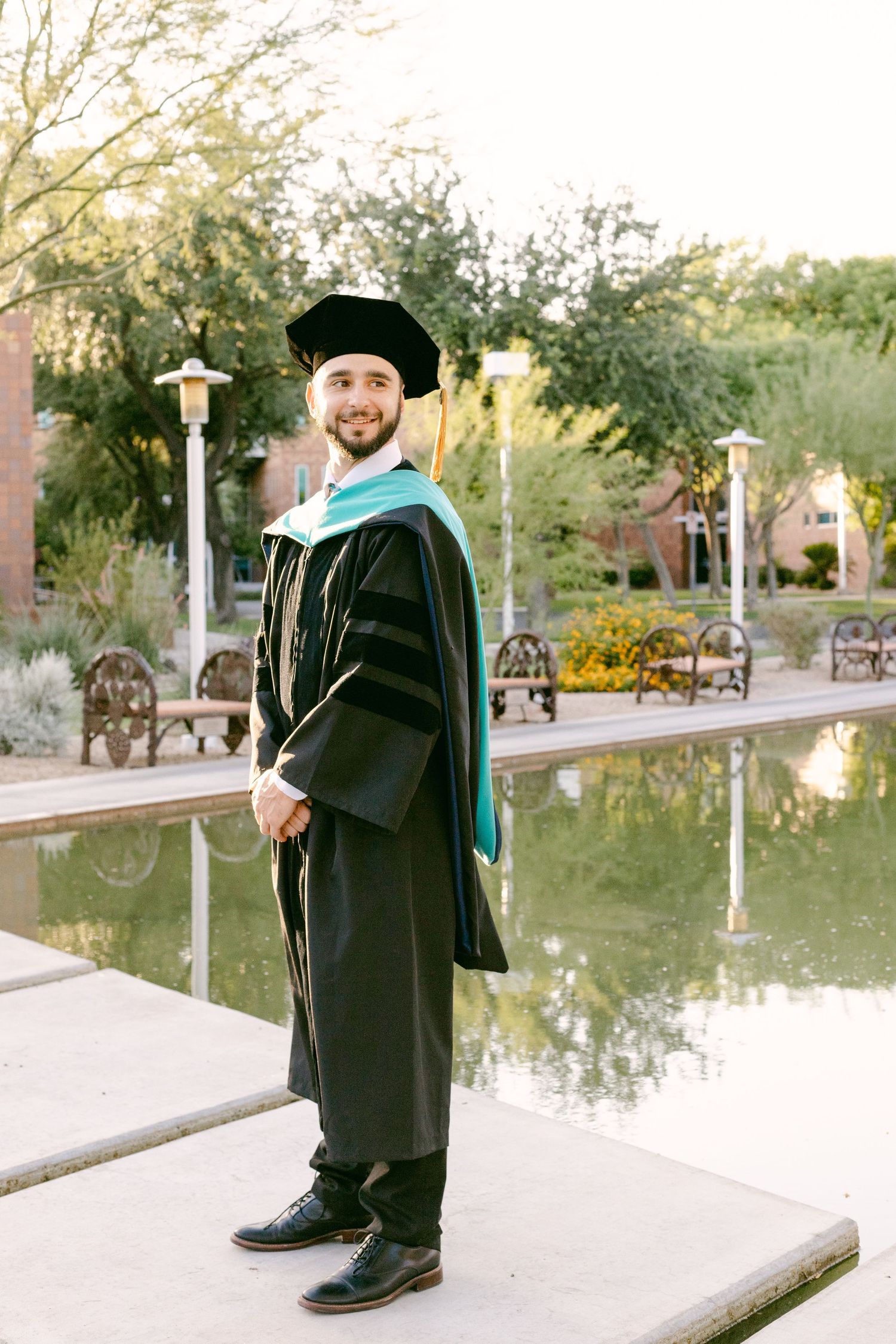 A master's graduate has his hands in front of him and is looking behind him. There's a pond behind him.