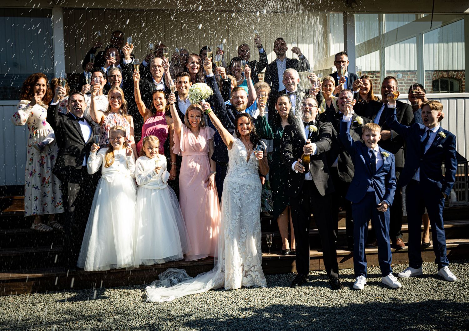 Large wedding party celebrating outdoors with confetti shower in front of a white building.