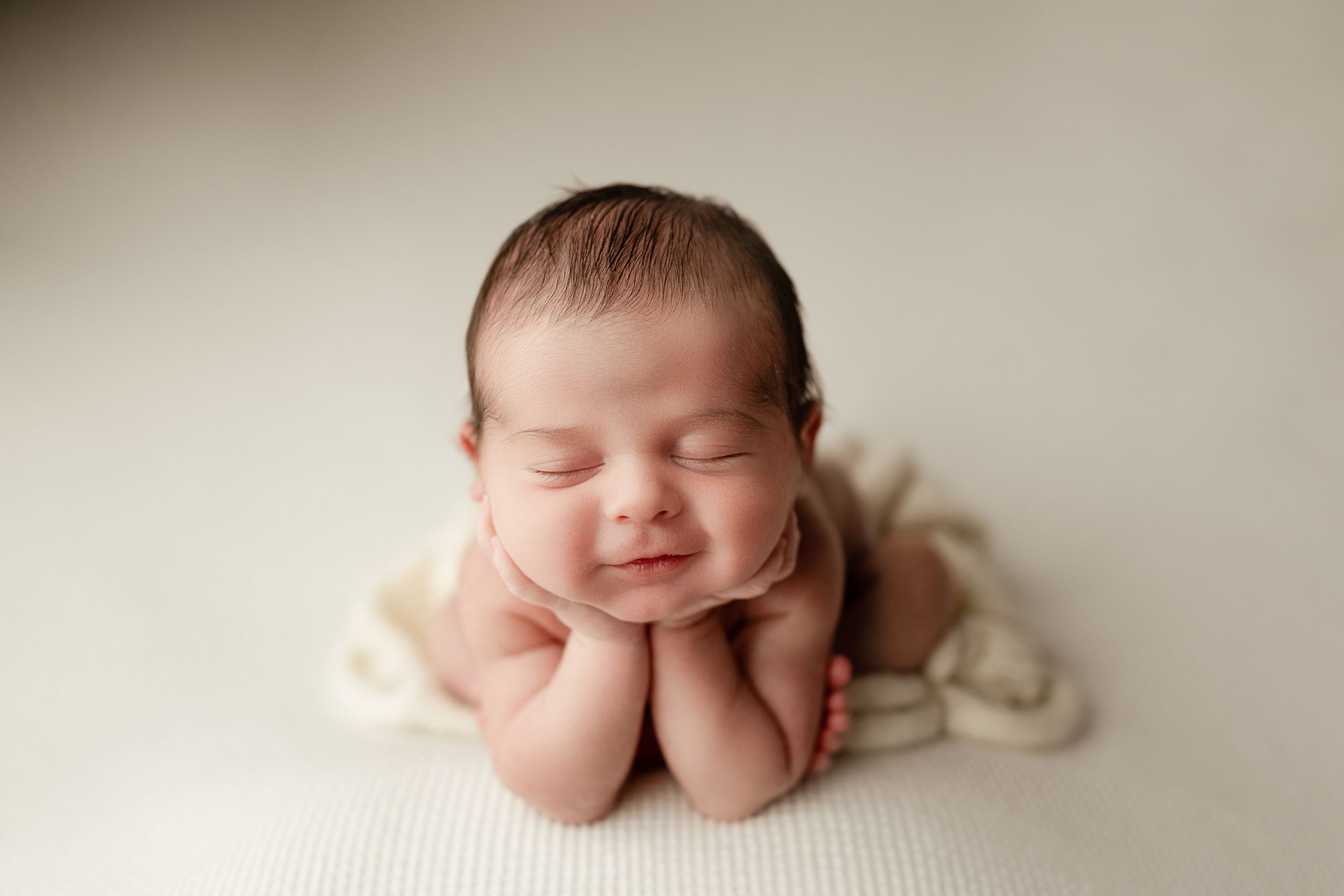 A sleeping newborn baby poses with hands under chin on cream-colored backdrop in studio lighting.