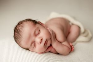 A peaceful sleeping newborn baby lies curled on a soft white fabric background.