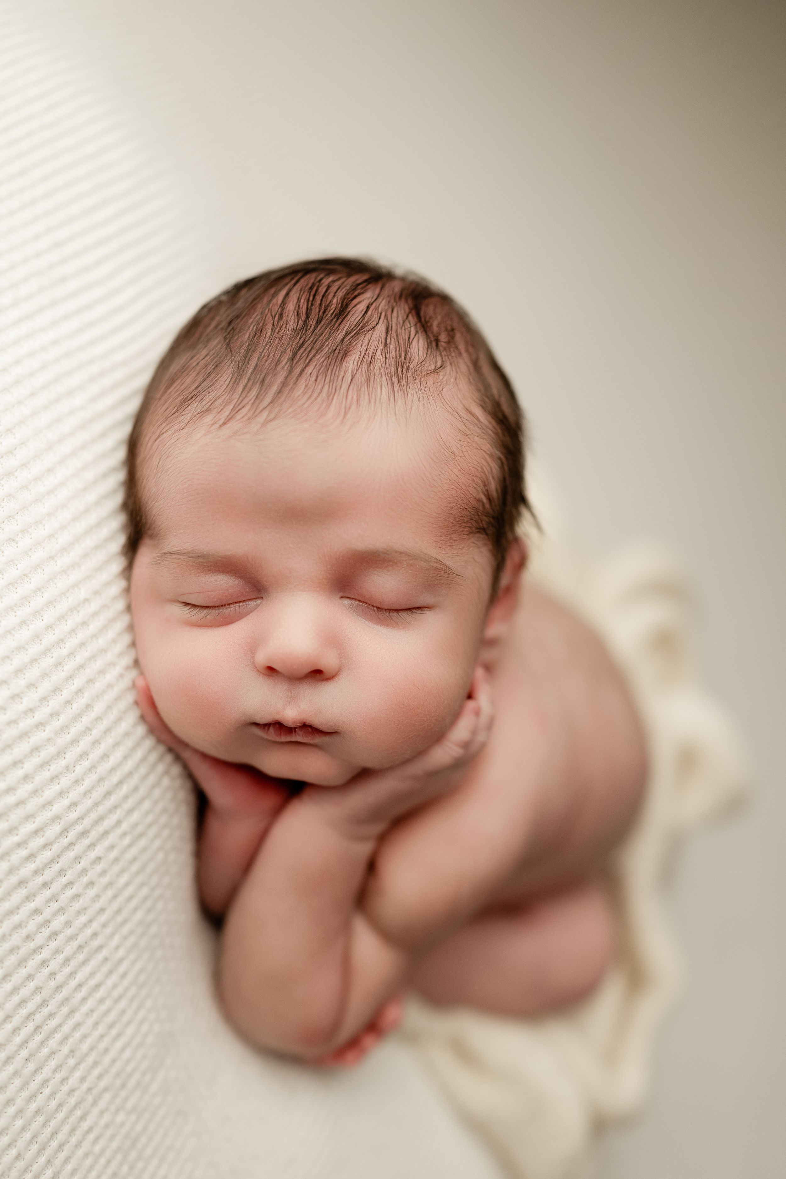 Sleeping newborn baby curled up peacefully on textured white fabric in soft lighting.