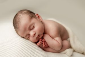 A peaceful sleeping newborn baby curled up on a cream-colored textured blanket.