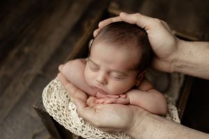 Peaceful sleeping newborn rests in supportive hands on a textured cream blanket against dark wooden background.