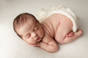 A peaceful sleeping newborn baby wearing a white diaper lies on textured white fabric.