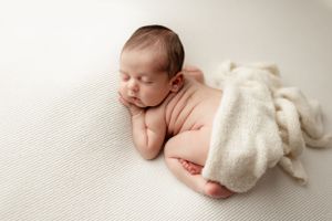 A peacefully sleeping newborn baby lies on white textured fabric with a soft cream blanket partially draped over.