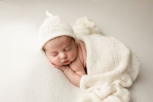 Sleeping newborn wearing white knitted hat and wrap on textured white background.