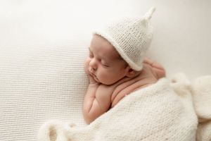 Peaceful newborn baby wearing knit hat resting on white textured blanket during photo session.