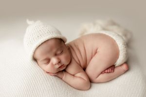Peaceful newborn in fetal position wearing white knit hat and diaper cover on white backdrop.