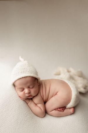 Peaceful sleeping newborn wearing white knit hat curled up on soft textured cream fabric.