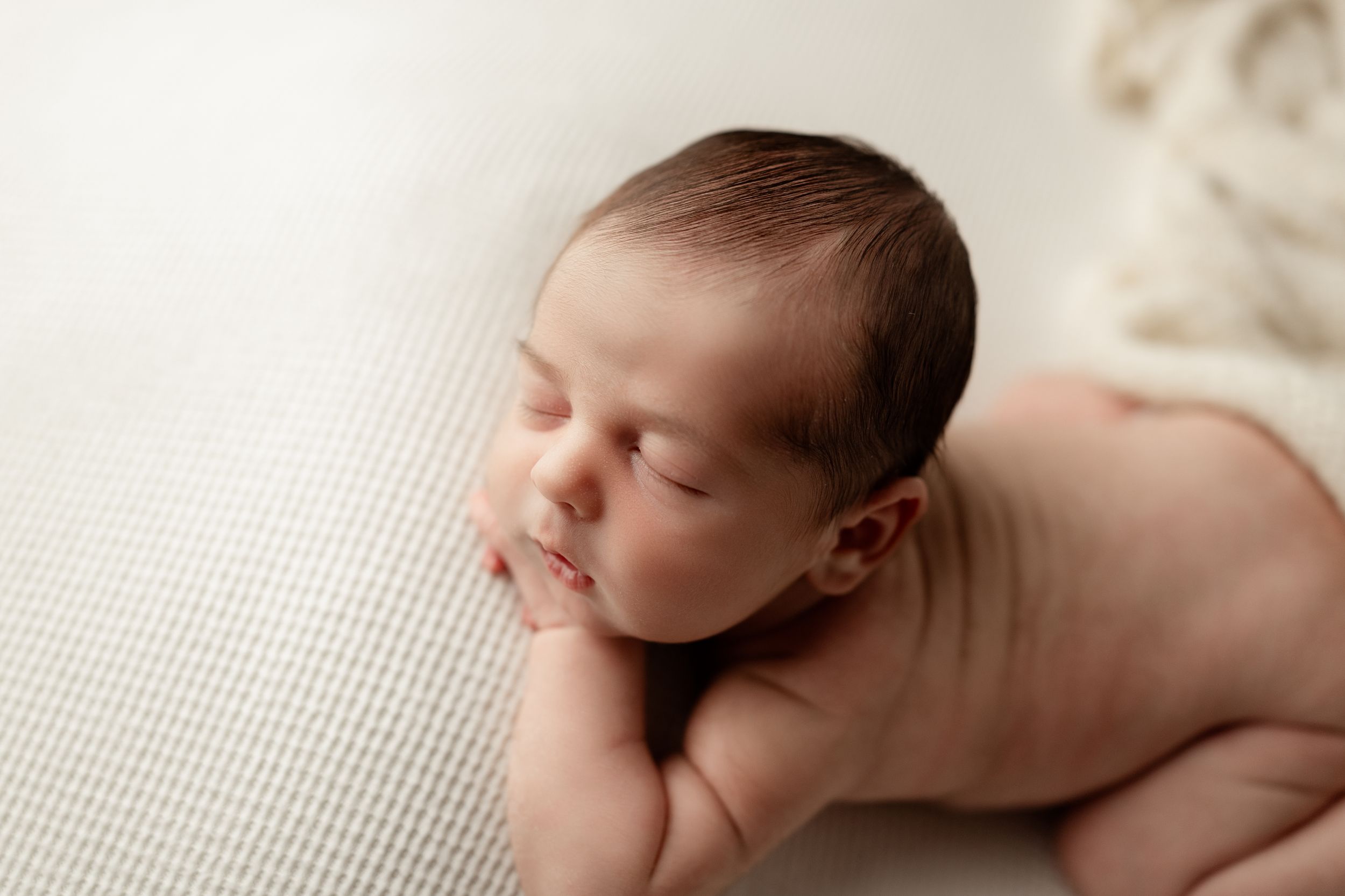 A sleeping newborn baby rests peacefully on a textured white blanket in a gentle side-lying pose.