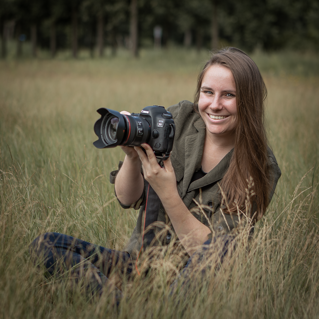 Fotografe zit in het gras met camera in de hand, lachend naar de lens.