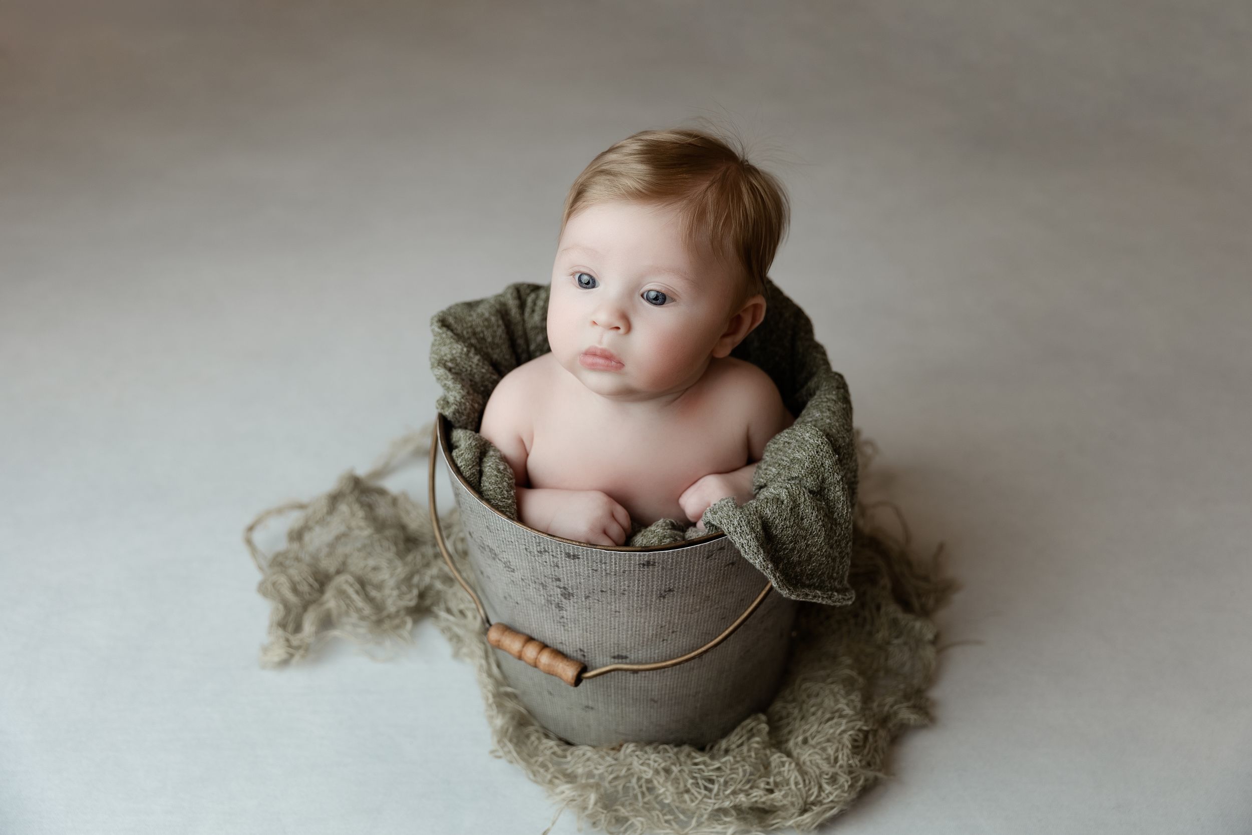 Baby sits in an olive green jacket on soft gray fur backdrop during minimalist studio photo session.
