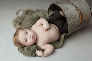 Baby lying in a metal bucket with an olive green blanket on a light gray background.