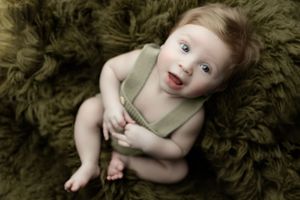 Baby wearing olive green romper lies on plush green rug while smiling with joy at camera.