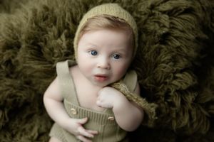 Baby wearing light green overalls lies on dark green fluffy textured backdrop.
