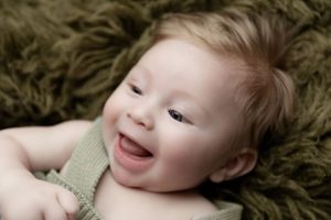 Joyful baby in sage green outfit smiles while resting on plush olive-colored surface.