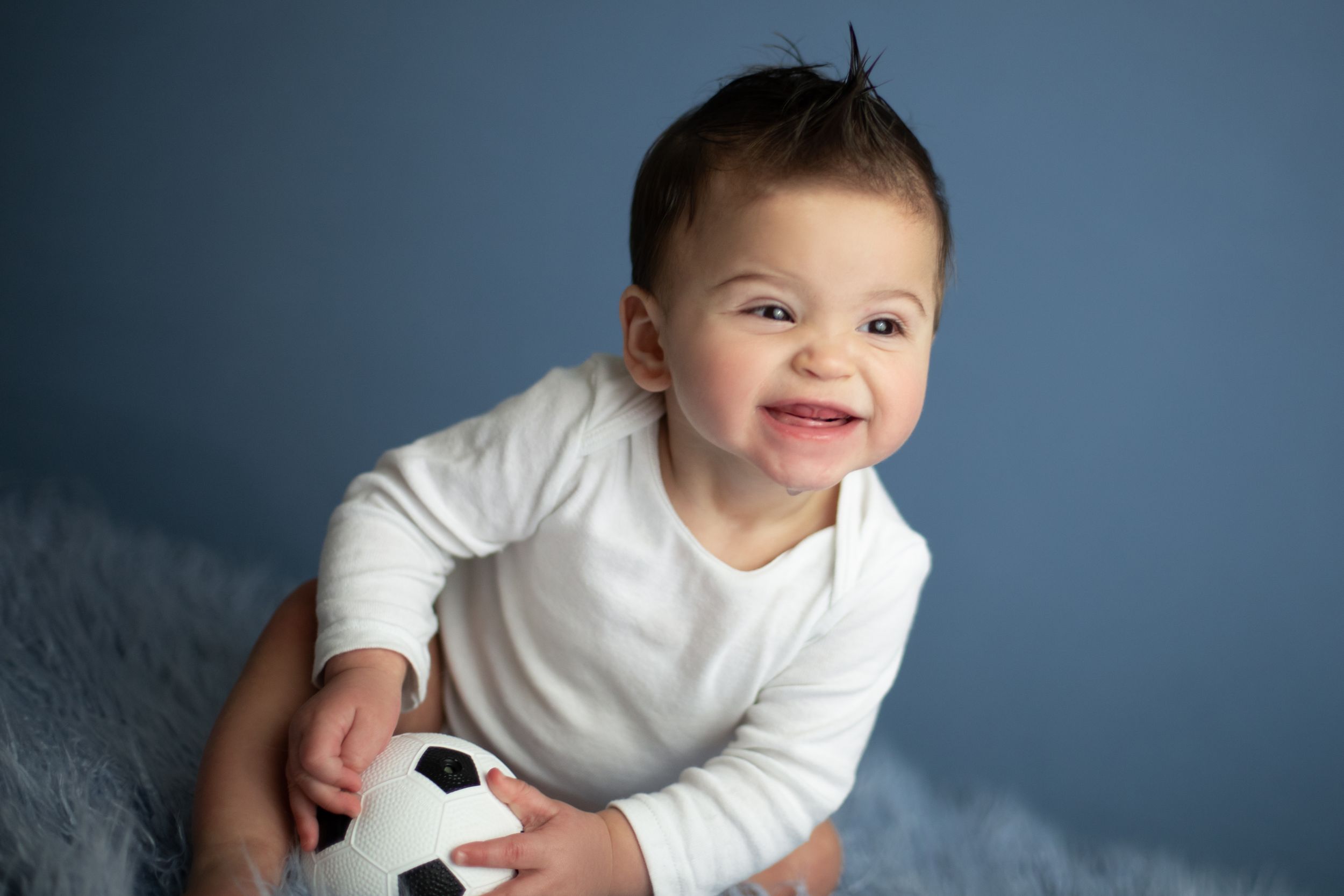 Smiling baby in white shirt holds soccer ball against blue background.