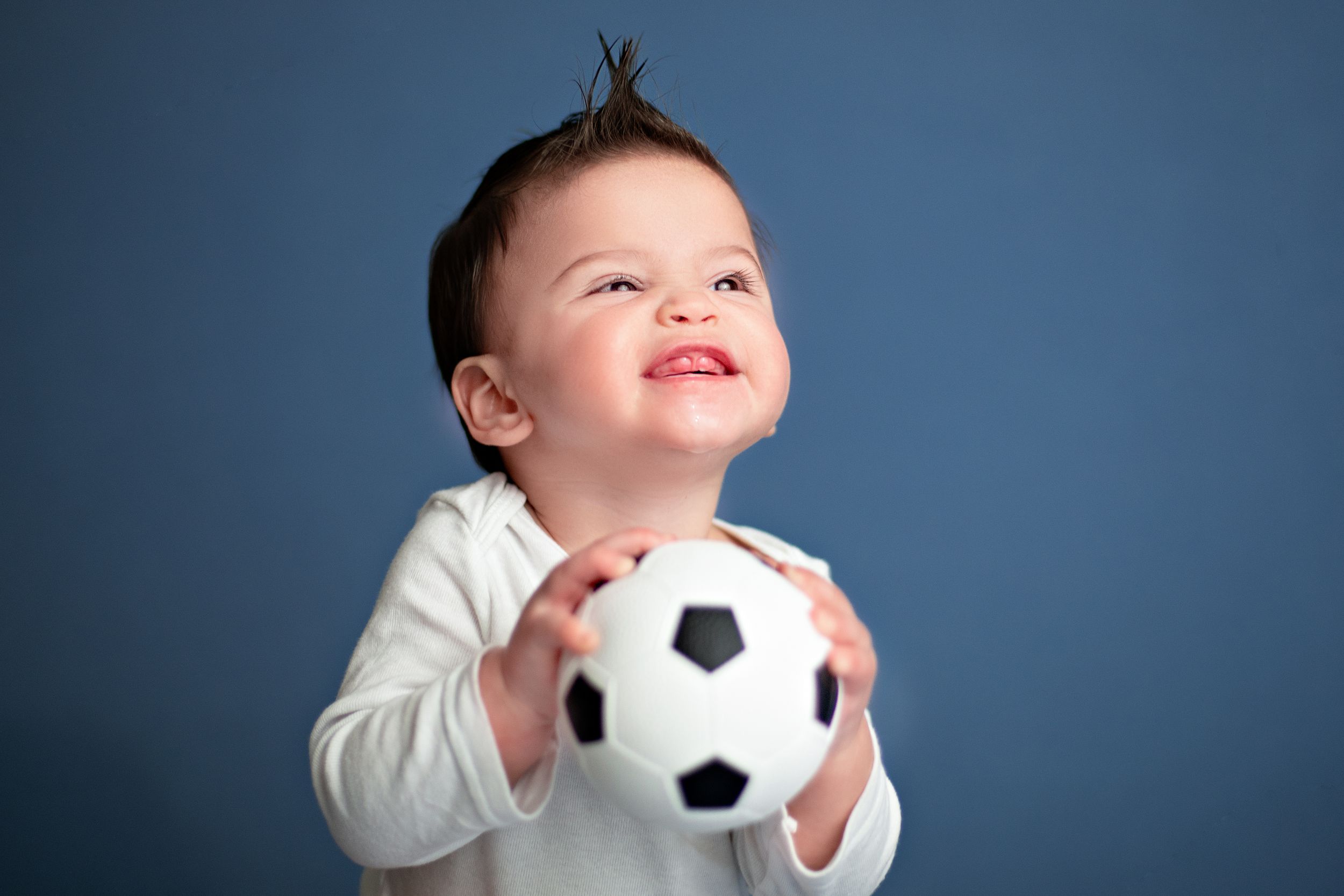 Child wearing white sweater holds a soccer ball while looking up with a happy expression against blue background.