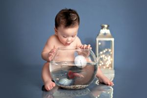 Baby reaches into glass fishbowl while sitting next to decorative lantern with fairy lights against blue background.