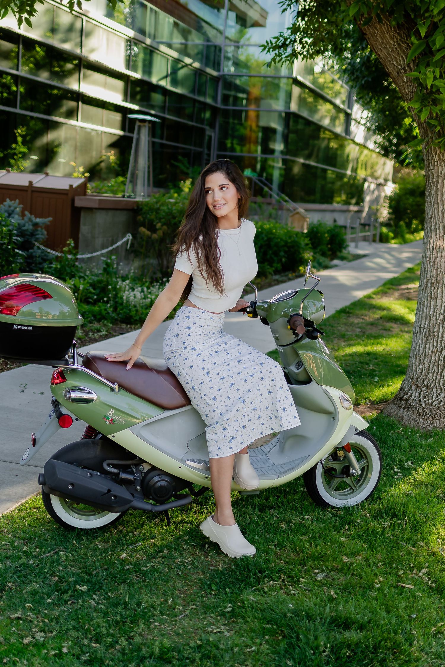 White scooter parked on grass near modern building with person wearing polka dot skirt and white top.
