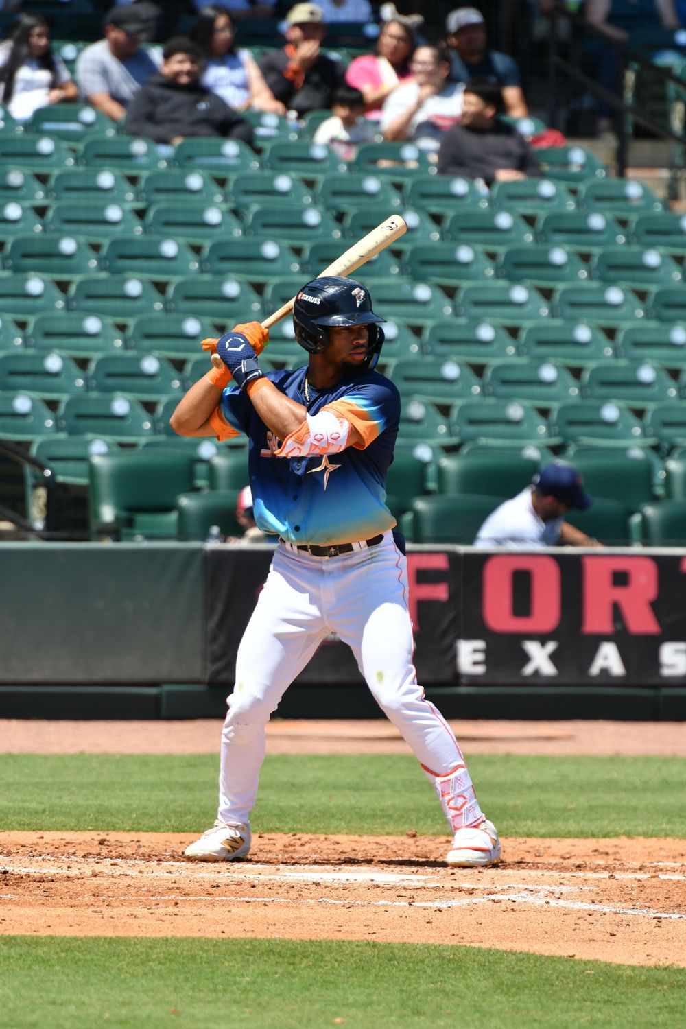 Baseball player in blue and white uniform stands at bat during daytime game with green stadium seats in background.