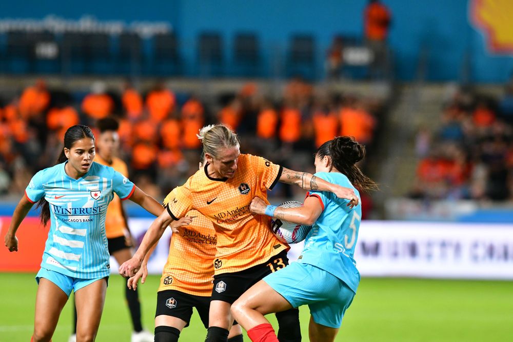 Soccer players in orange and light blue uniforms compete for ball possession during match.