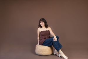 Portrait of person in brown top and jeans seated on woven ottoman against brown backdrop.