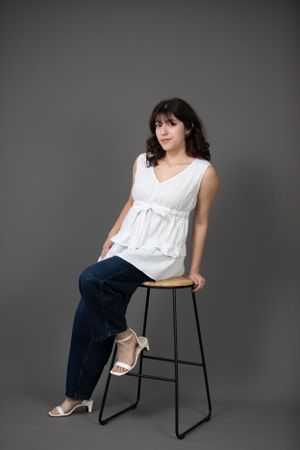 Studio portrait with white flowing top and jeans seated on black metal stool.