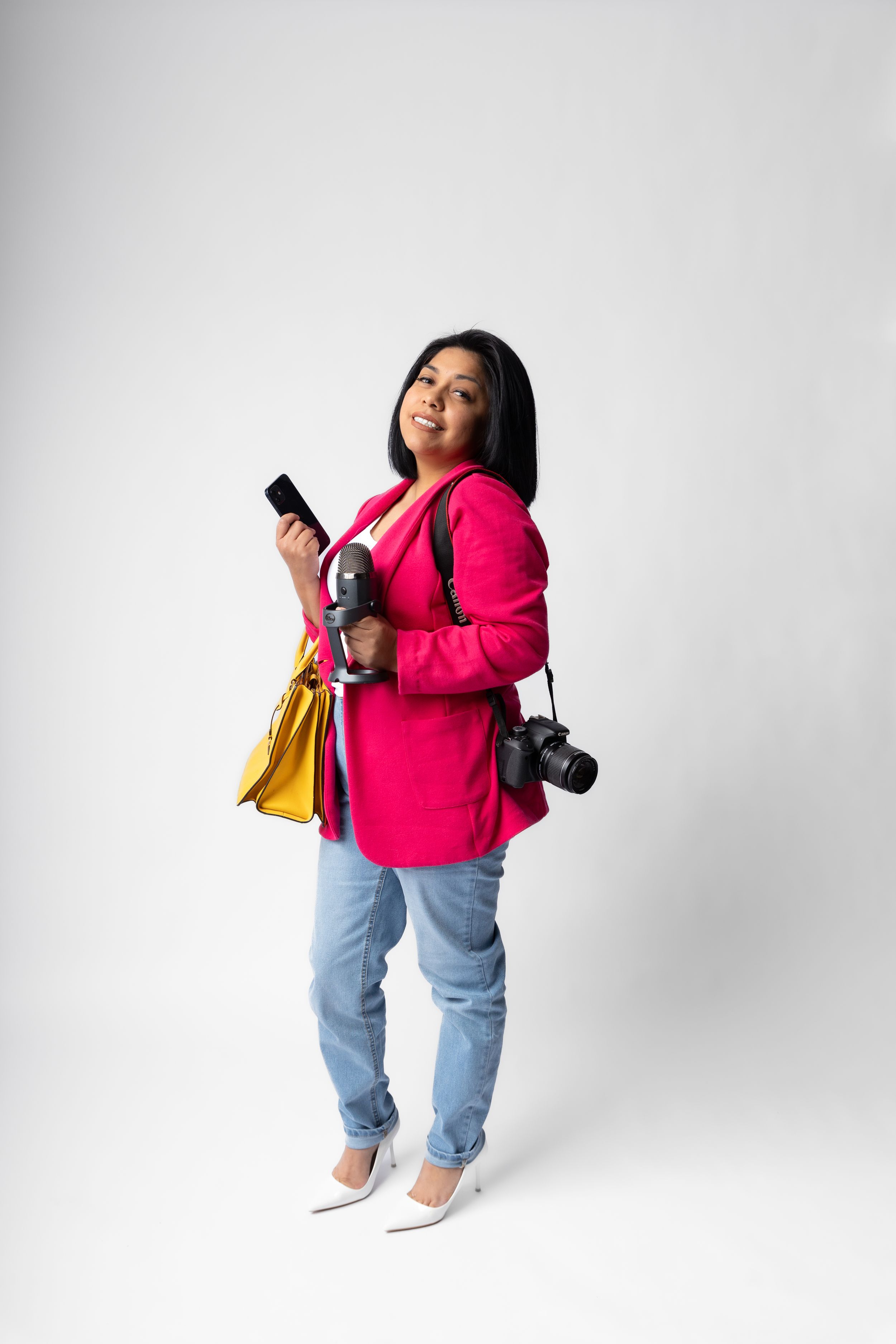 Person in bright pink blazer and jeans holding phone and camera with yellow bag against white background.