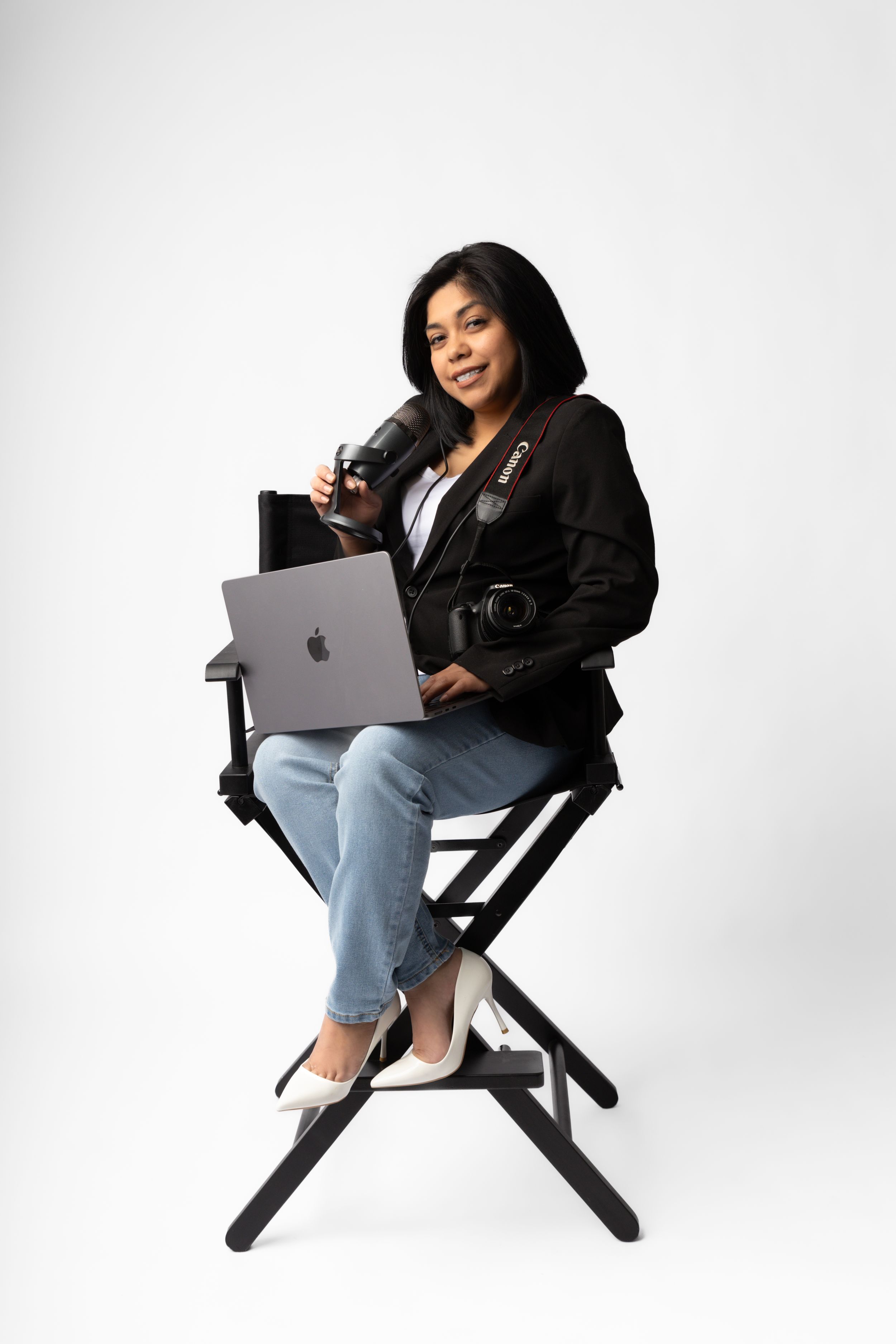 Professional photographer sits in director's chair with laptop and camera equipment on white background.