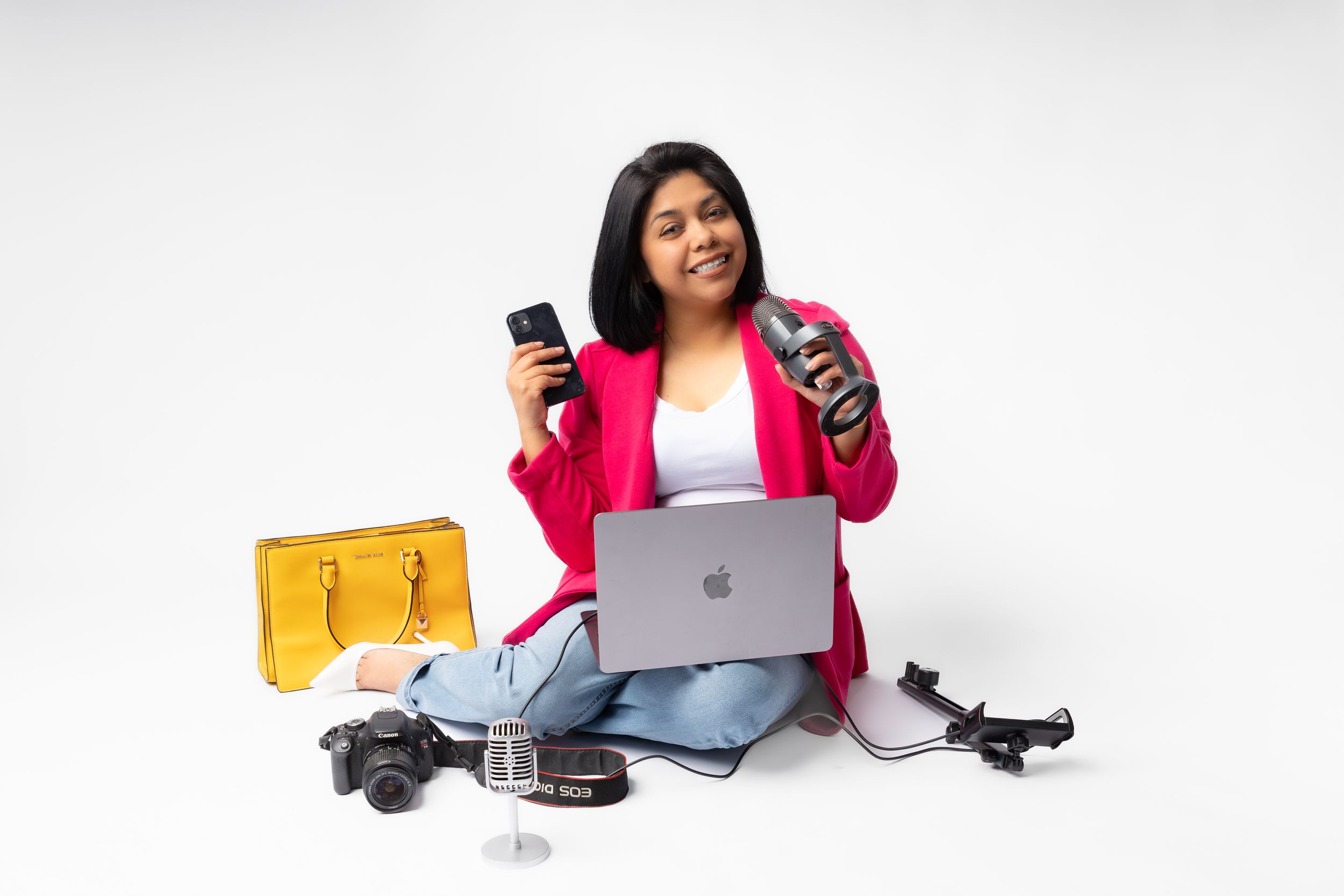 Content creator sits on floor with laptop, cameras, and yellow handbag against white background.