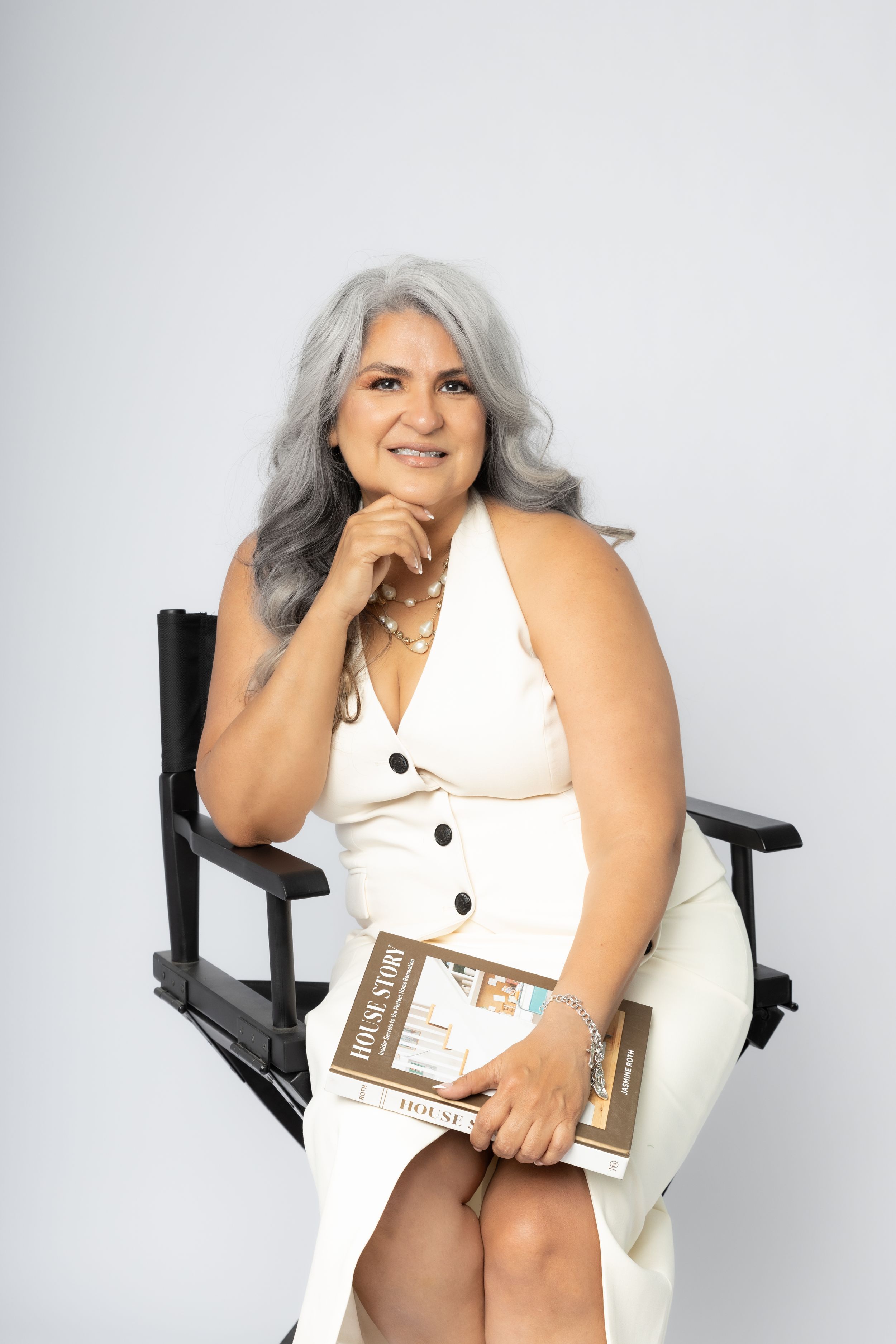 Plus-size model in white sleeveless dress sitting confidently in director's chair against light background.