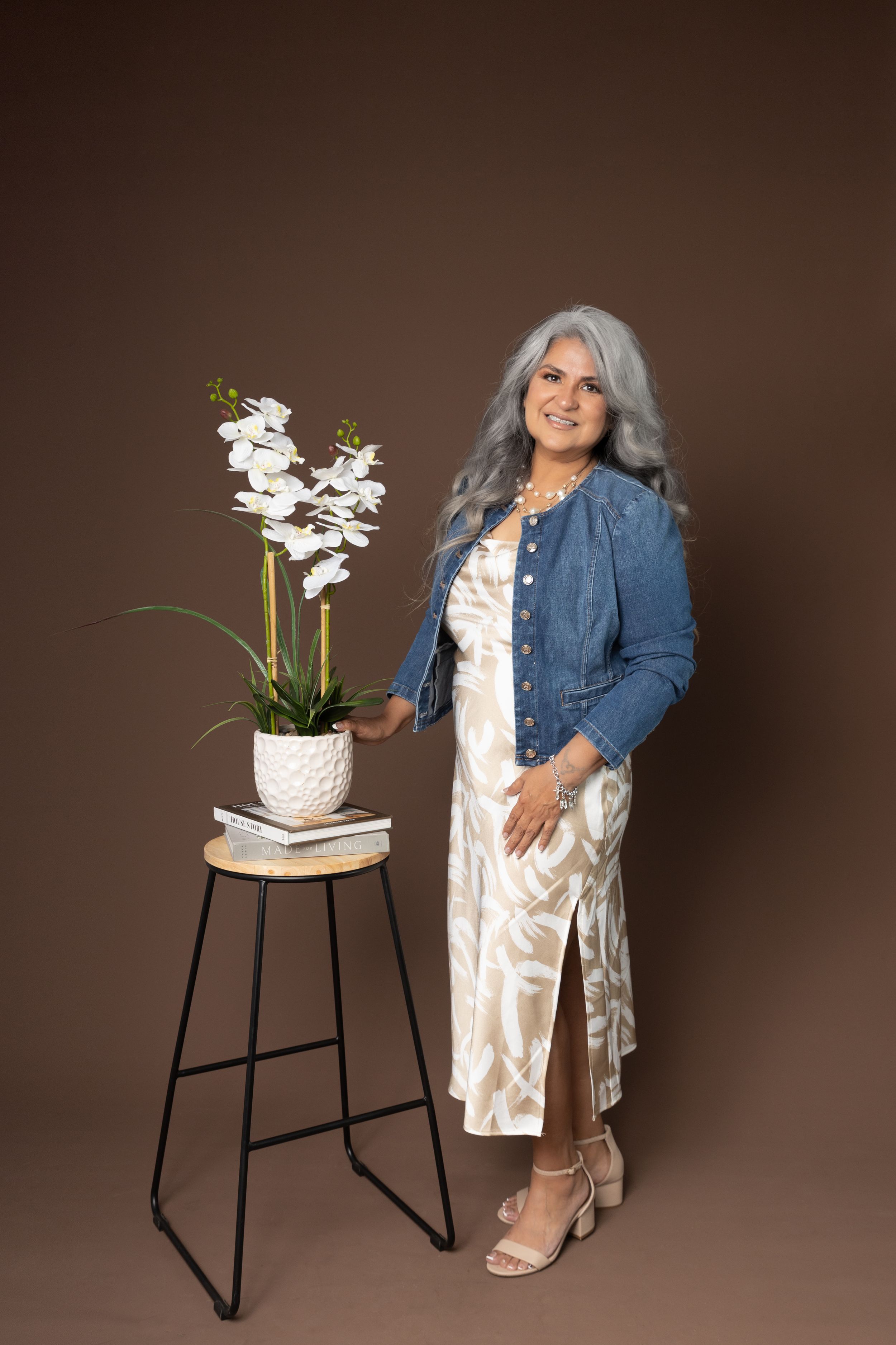 Stylish studio portrait with a blue denim jacket, floral dress, and decorative white orchid on metal stool against brown backdrop.