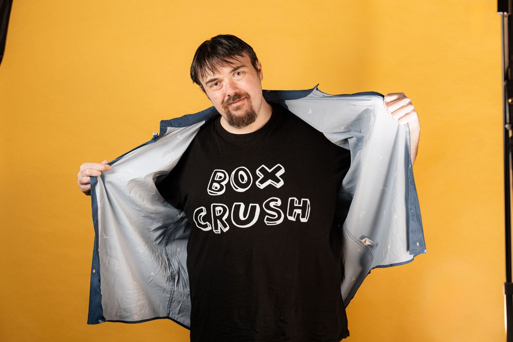 A person wearing a black 'Box Crush' t-shirt poses against a yellow background while holding open a gray jacket.
